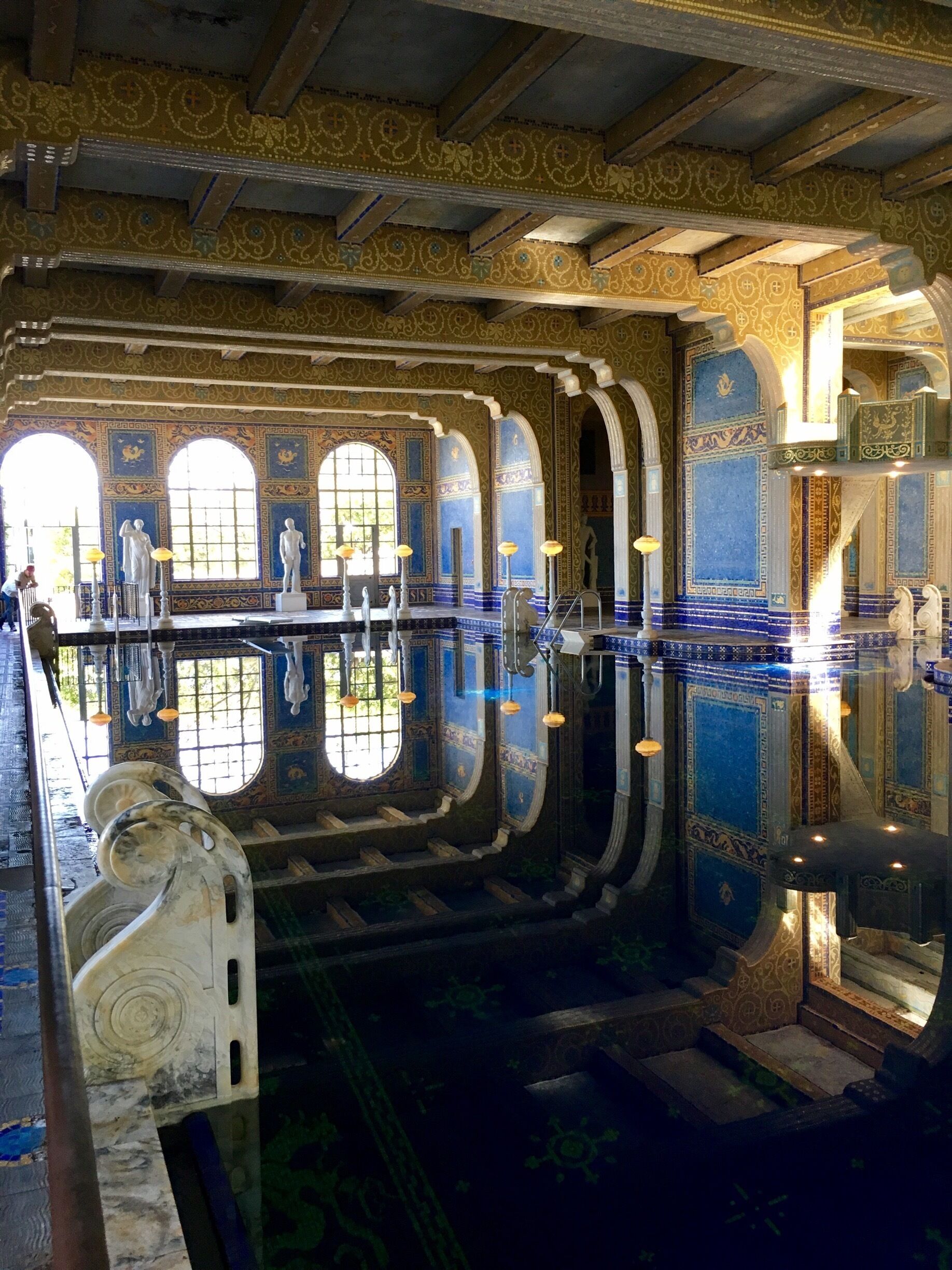 A grand indoor pool with ornate blue and gold tile work, large arched windows, and classical statues.