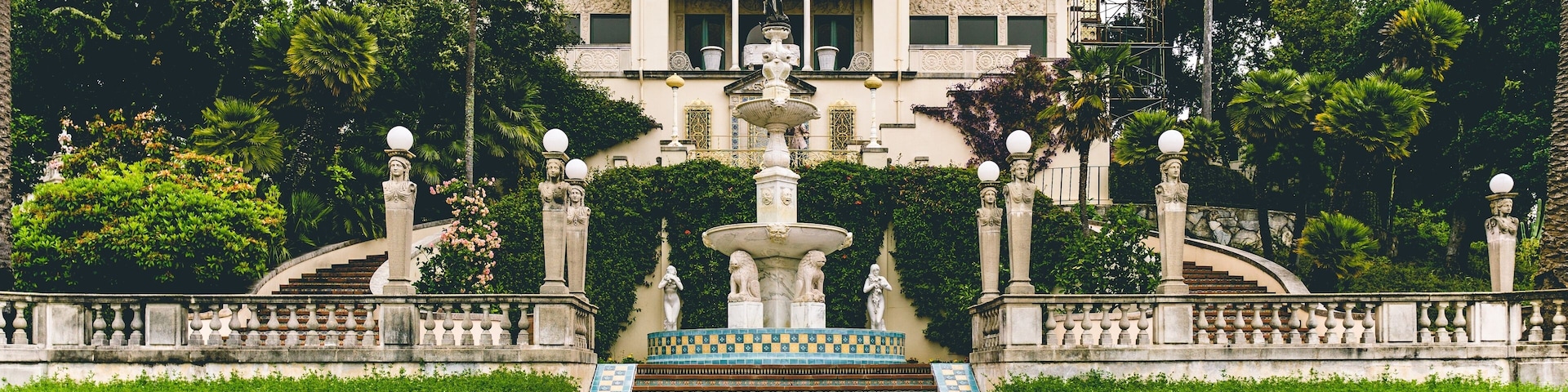 A grand building with a central fountain, surrounded by lush greenery and palm trees.