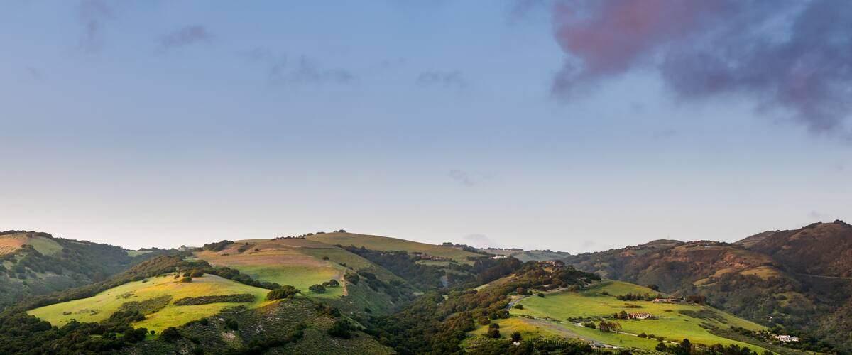 Panorama of beautiful green hills, mountains, and fields in sunset light under a blue sky with purple clouds in Carmel Valley, CA, USA