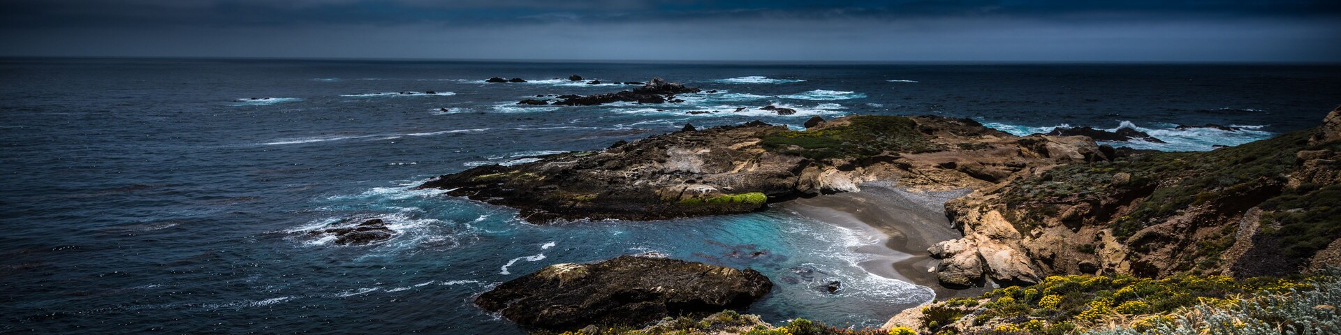 Paisagem costeira com falésias rochosas, uma praia arenosa e um mar agitado sob um céu nublado.