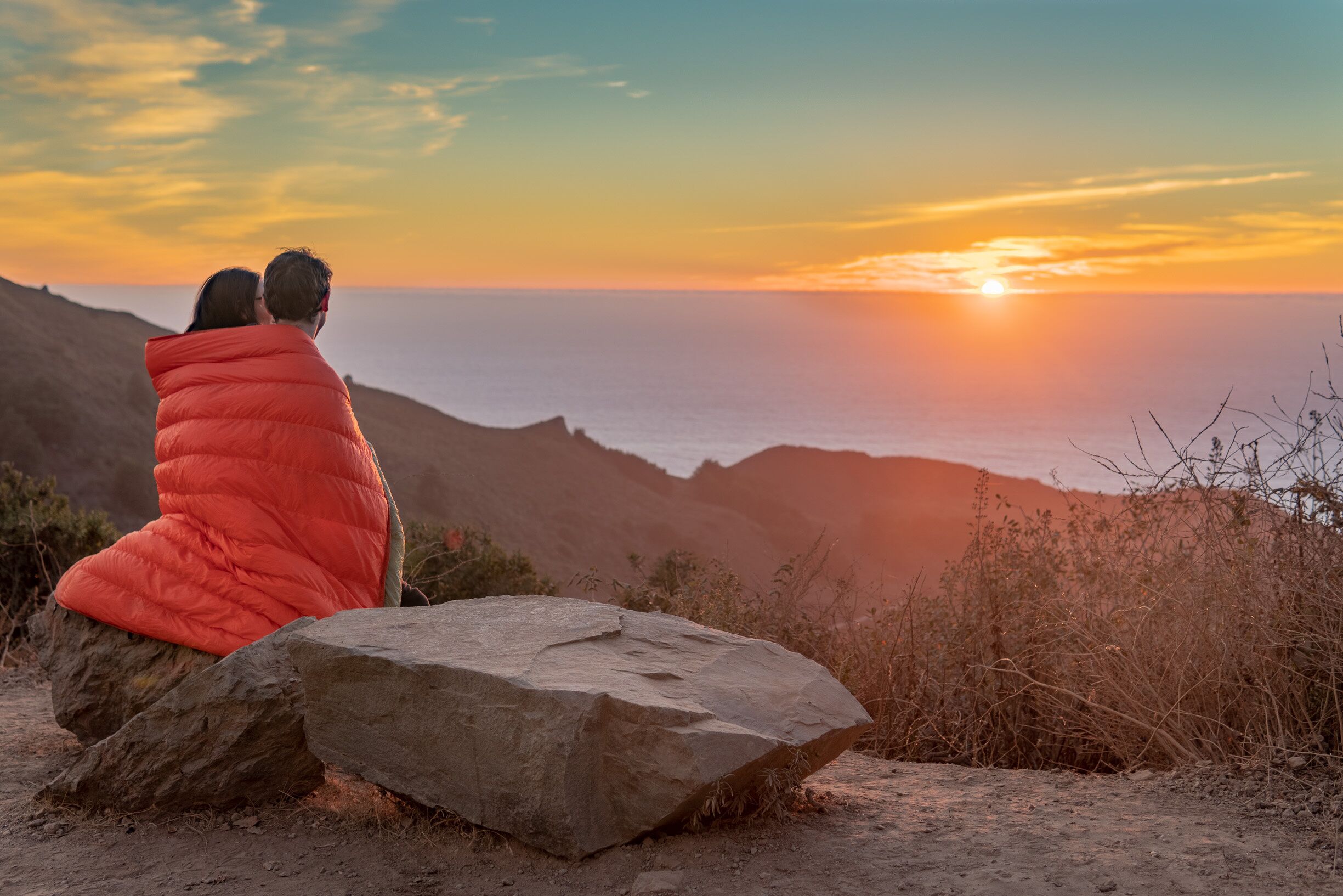 Two people sitting on a rock overlooking a sunset over a mountainous landscape.