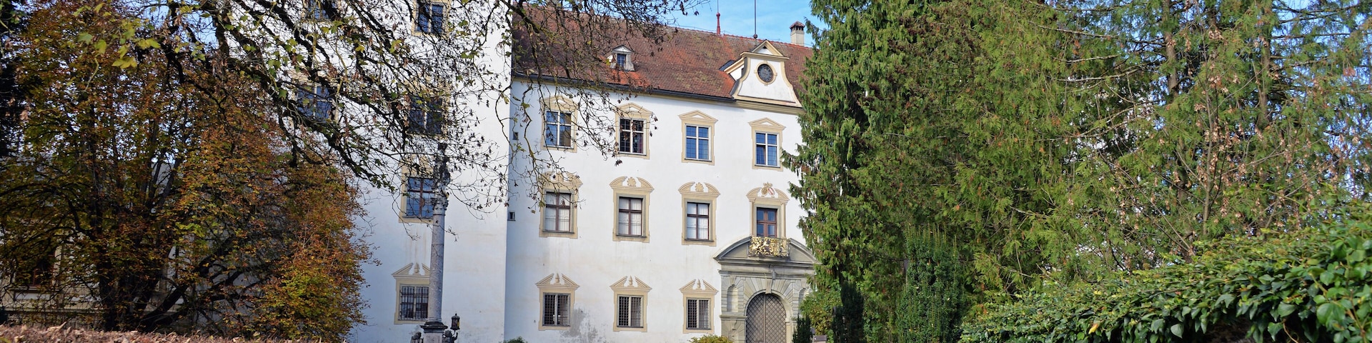 Um edifício histórico com fachada branca e telhado de telhas vermelhas, rodeado por árvores e um portão de metal.
