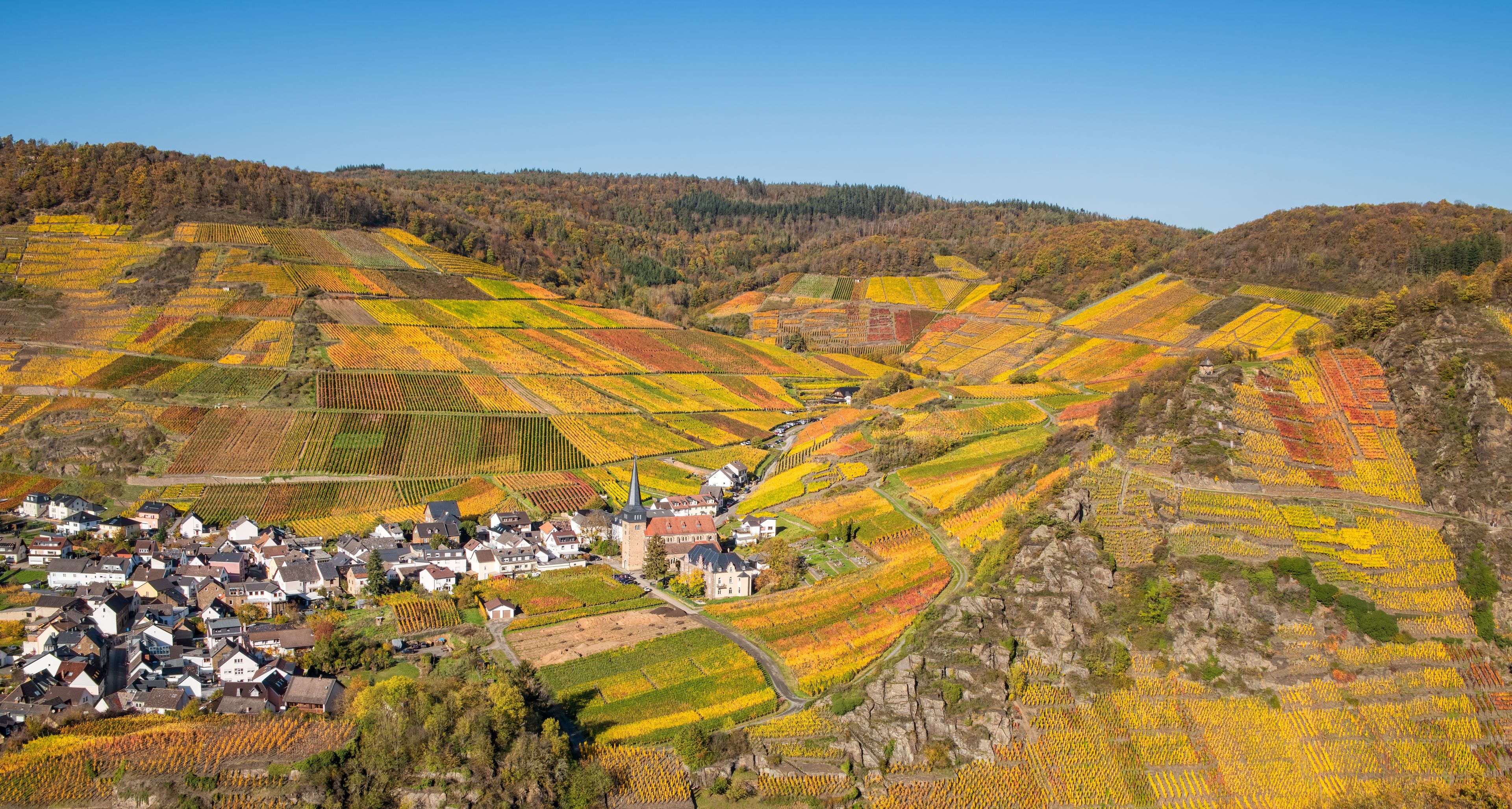 A village nestled in a valley with terraced vineyards and a church with a tall spire.