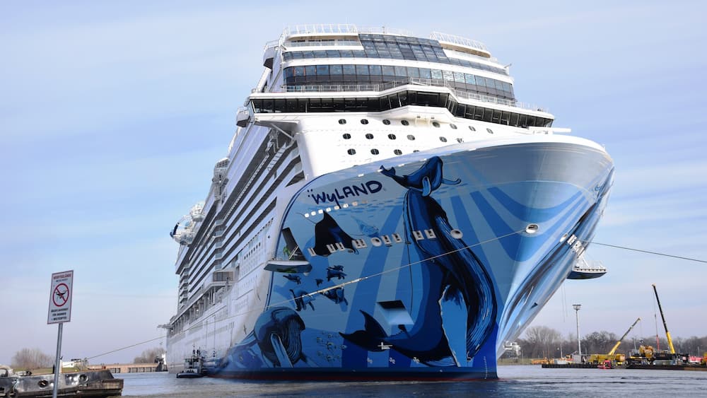 Ein großes Kreuzfahrtschiff mit der Aufschrift 'WYLAND' an der Seite, liegt im Hafen an, wo sich ebenfalls Kräne und ein Schlepper befinden.