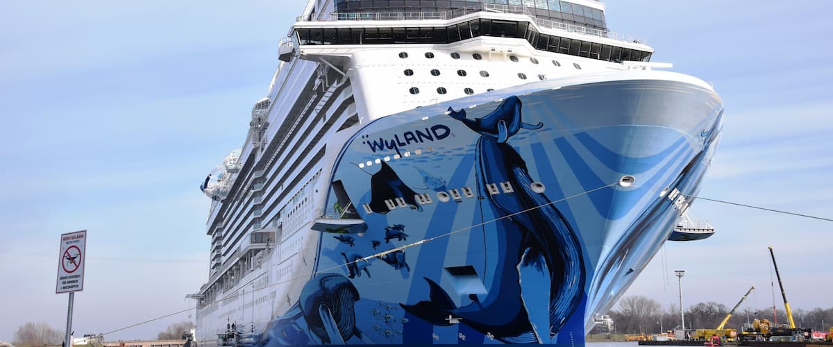 Un gran crucero con la leyenda 'WYLAND' en su lateral, atracado en un puerto con grúas y un remolcador cerca.