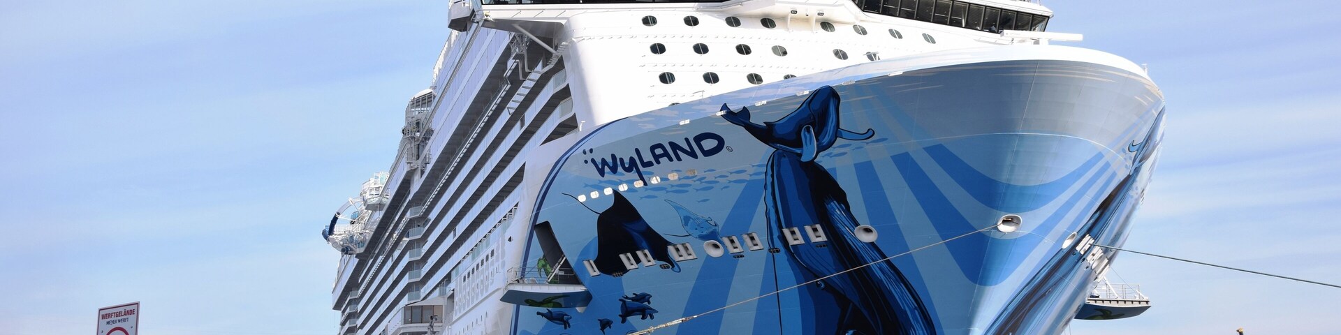 Un gran crucero con la leyenda 'WYLAND' en su lateral, atracado en un puerto con grúas y un remolcador cerca.