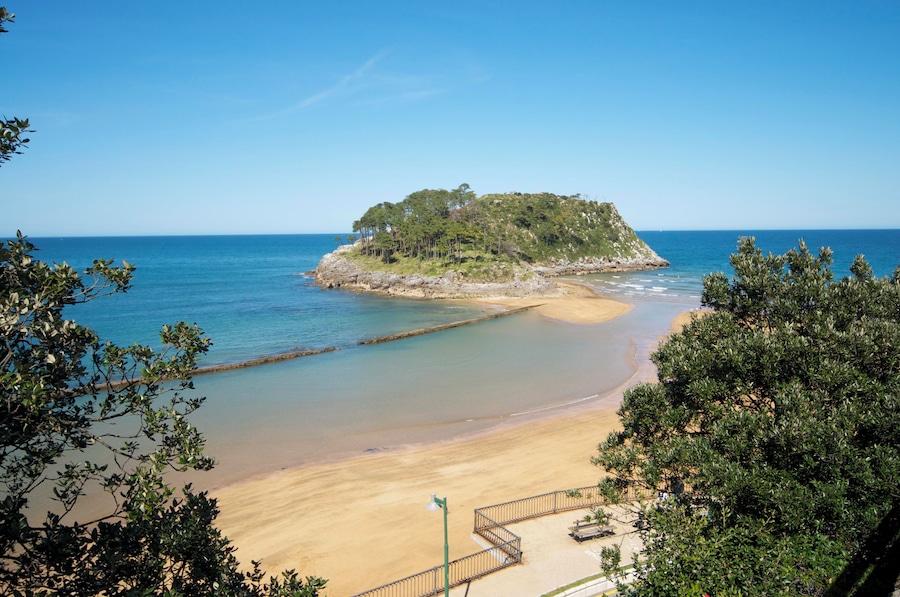 A beach with a small island in the distance, surrounded by clear blue water and green foliage.