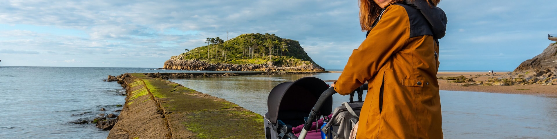 A coastal landscape with a rocky shoreline, a small island in the distance, and a person with a stroller.
