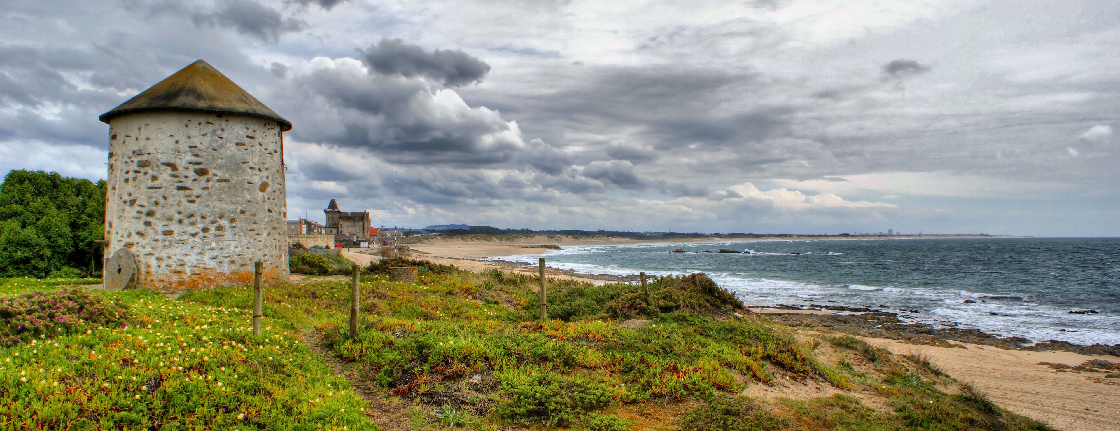 A coastal landscape with a stone tower, beach, and ocean under a cloudy sky.