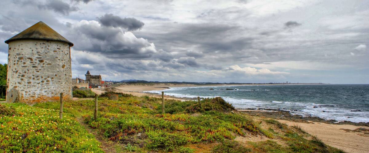 A coastal landscape with a stone tower, beach, and ocean under a cloudy sky.
