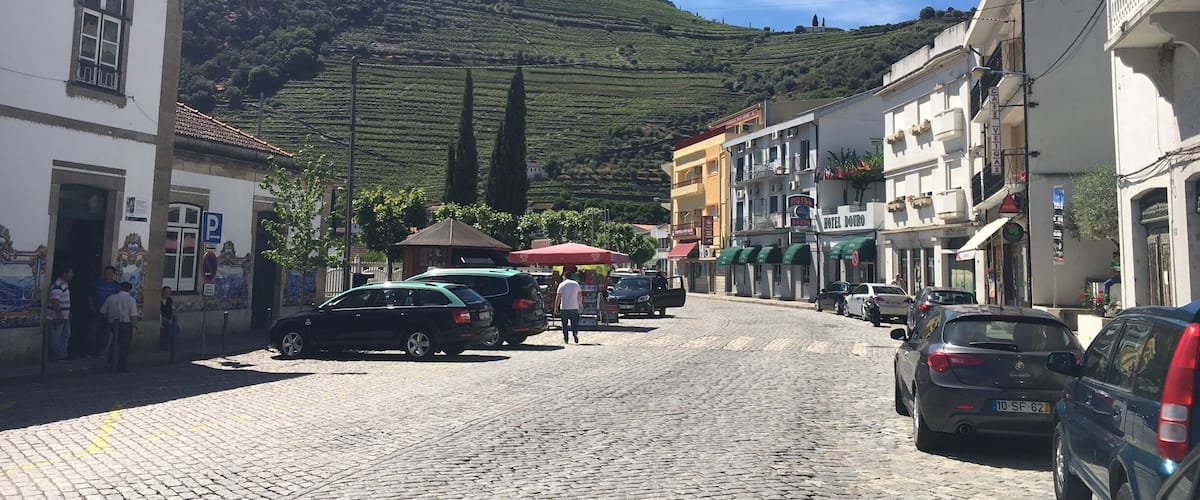 A cobblestone street lined with buildings, parked cars, and a hill with vineyards in the background.