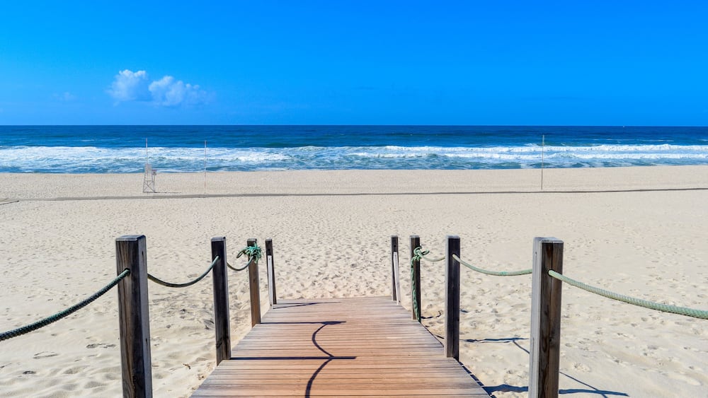 Ein hölzerner Steg führt zu einem Strand mit blauem Himmel und Meereswellen.