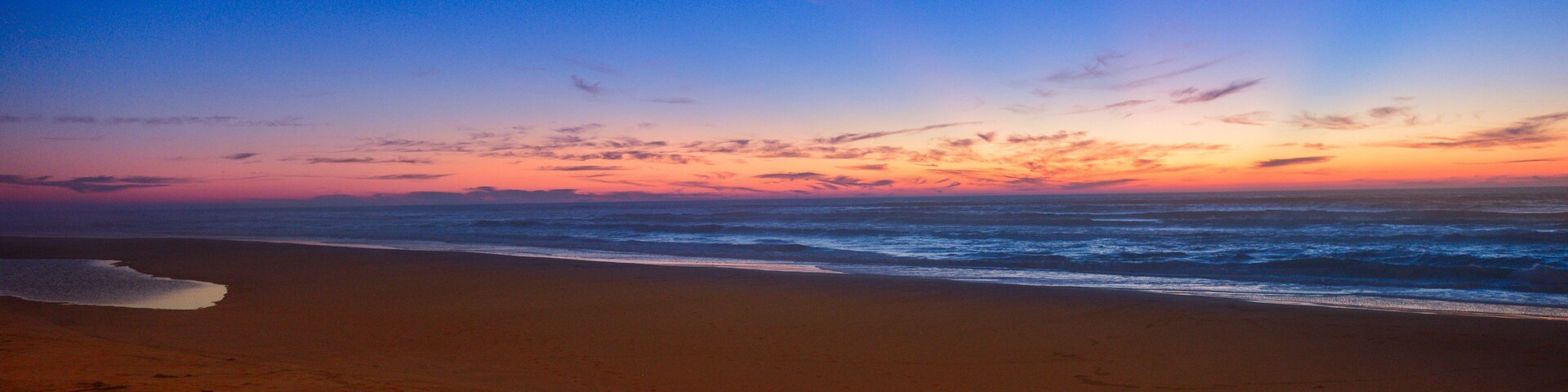 Uma praia ao pôr do sol com ondas, céu claro e pegadas na areia.