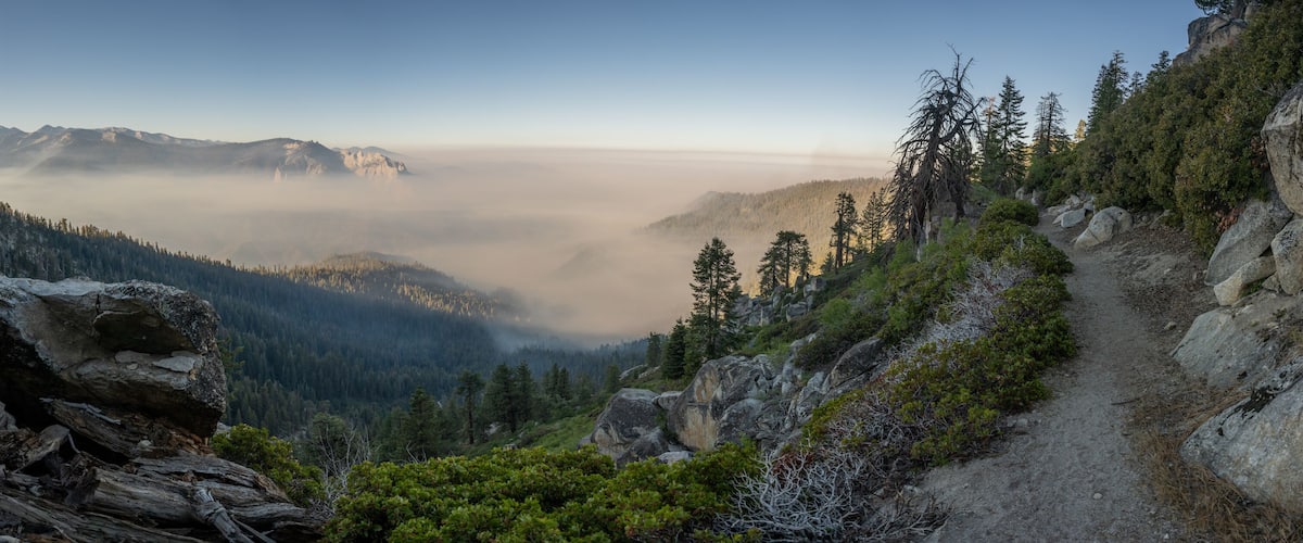 A mountainous landscape with a forested valley, a clear sky, and a rocky path.