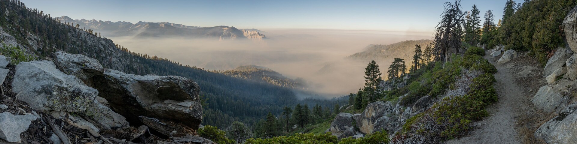 A mountainous landscape with a forested valley, a clear sky, and a rocky path.