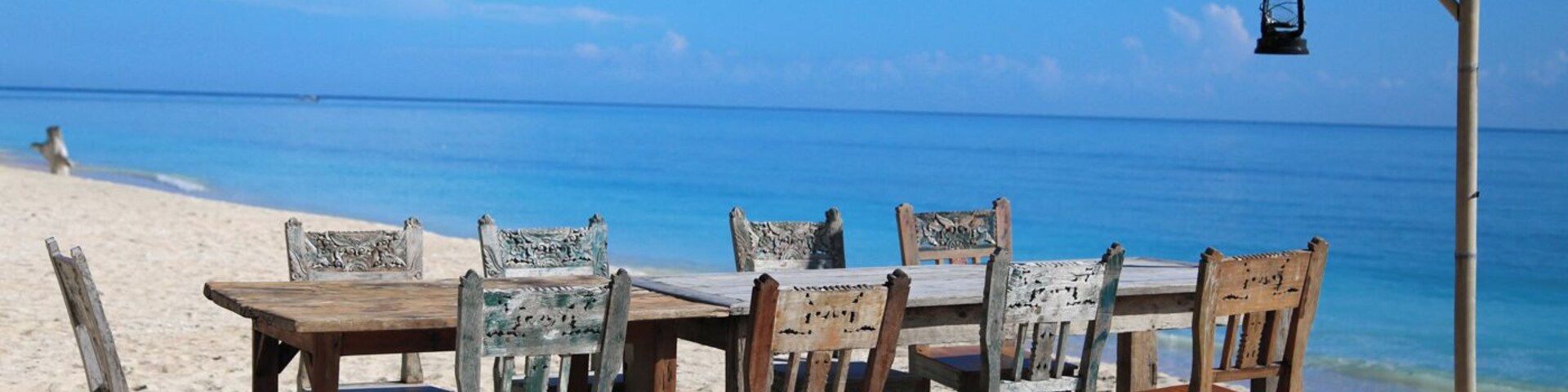 Una mesa y sillas de madera sobre una playa arenosa, con un cielo azul despejado y el océano al fondo.