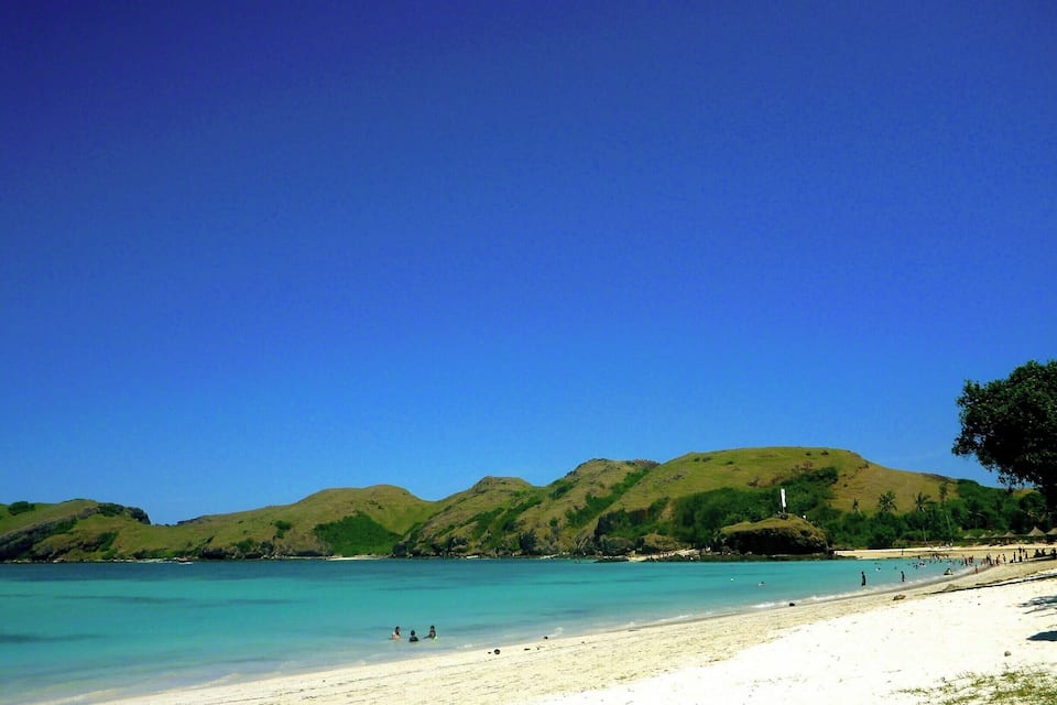 Une plage avec une eau turquoise cristalline, du sable blanc et des collines verdoyantes en arrière-plan.