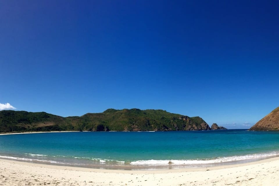 Una playa con agua turquesa cristalina, arena blanca y colinas verdes al fondo.