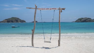 Um parquinho de baloiço de madeira numa praia arenosa, com uma placa a indicar "Tanjung Arau Lombok" e um barco na água.
