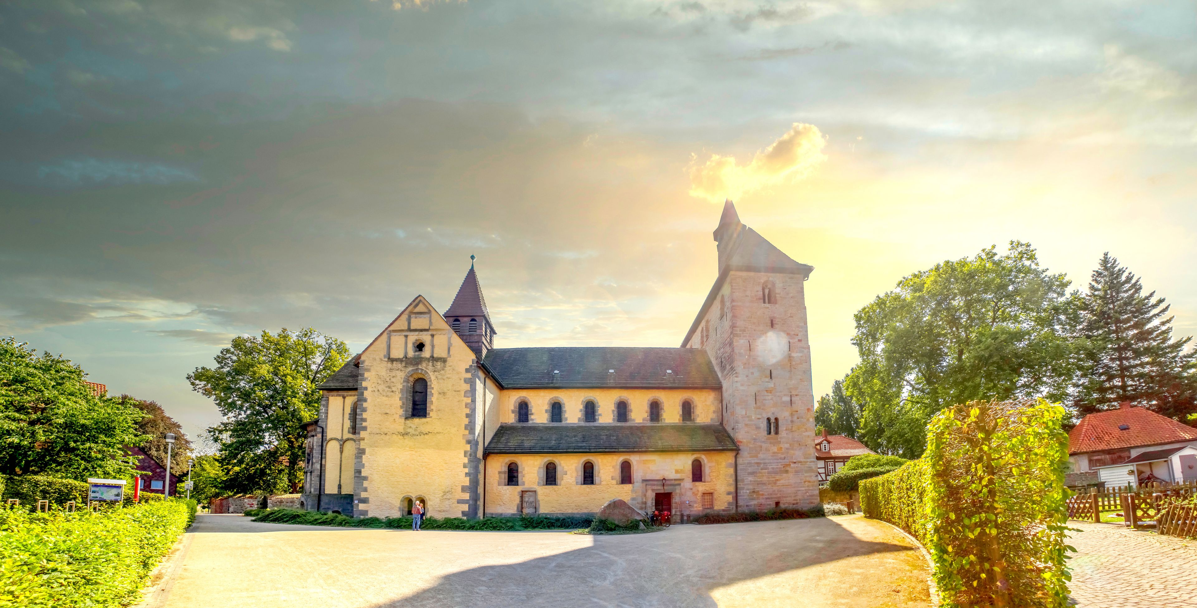 A historic church with a prominent tower and a smaller adjacent building, surrounded by lush greenery and a paved driveway.