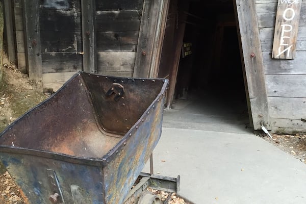 A rusty metal wheelbarrow in front of a wooden building labeled "Gold Bug Mine" with a sign that says "OPEN".