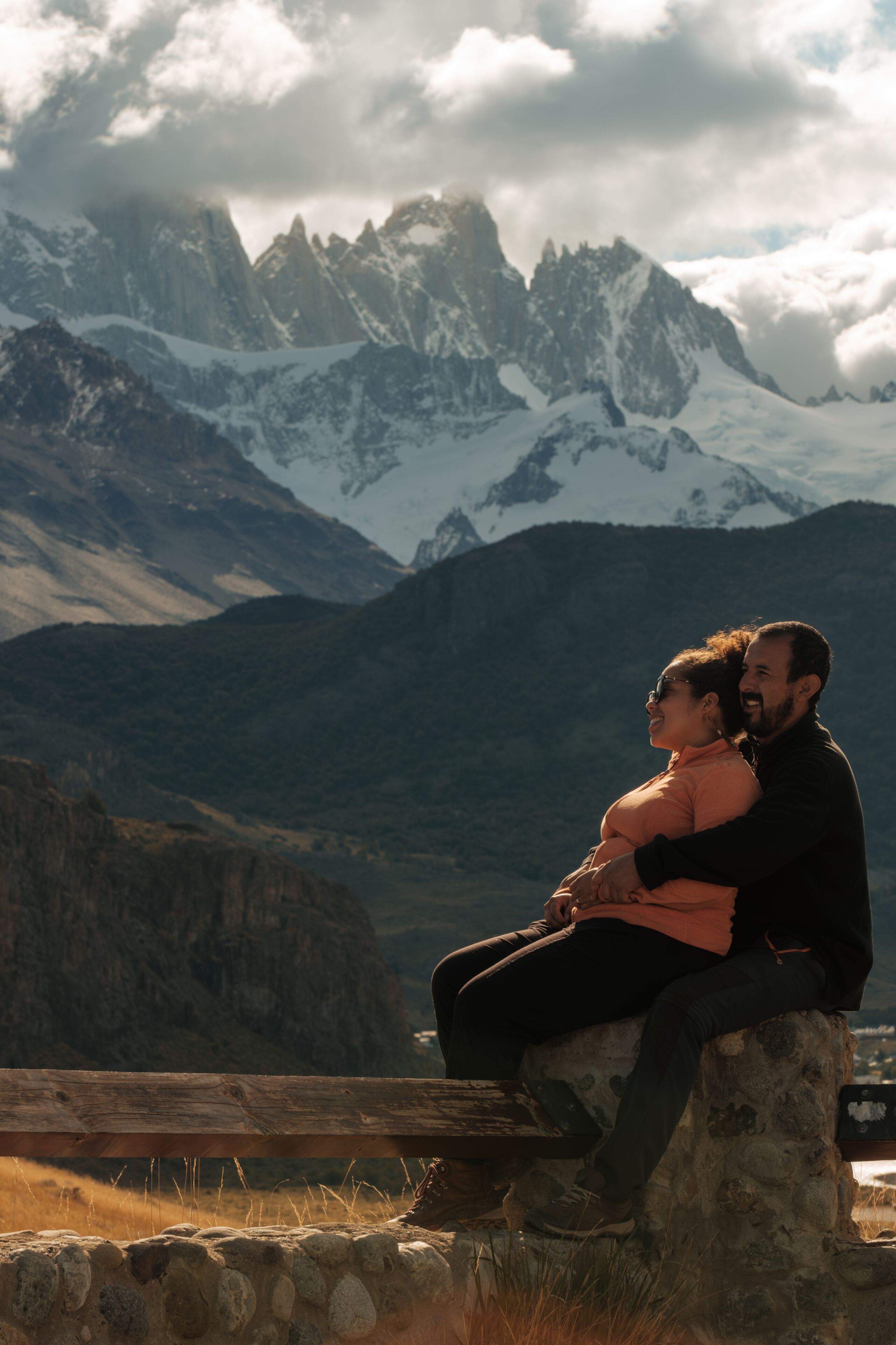Una pareja sentada sobre un muro de piedra con vistas a un paisaje montañoso con picos nevados.