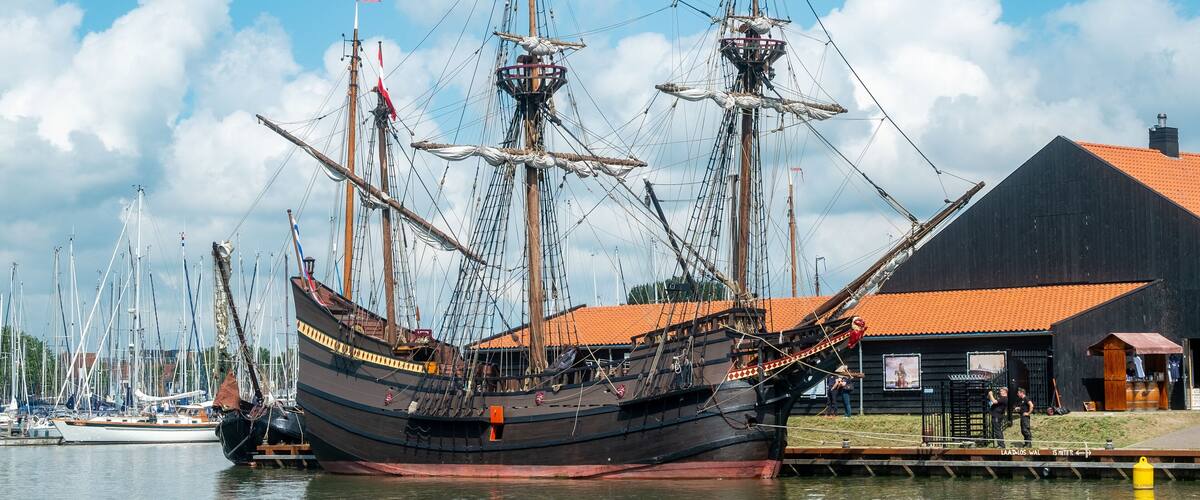 A large sailing ship docked at a marina with a black building and a red-roofed structure nearby.
