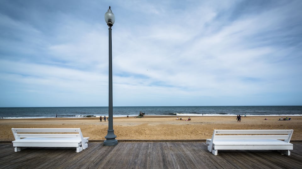 A wooden boardwalk with two white benches and a street lamp overlooking a beach with people and a boat.