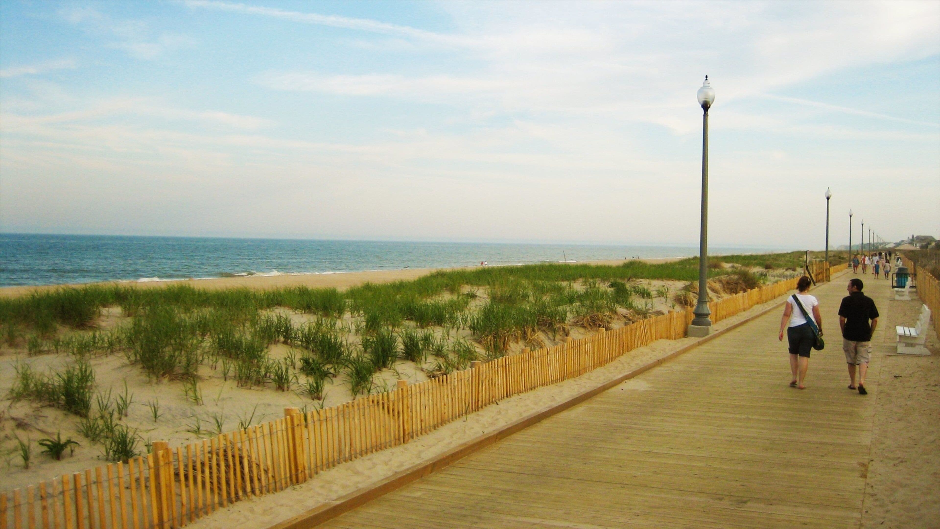 A wooden boardwalk along a beach with dunes and a fence.