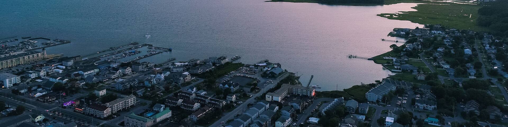 An aerial view of a coastal town at sunset with a large body of water, a pier, and residential areas.