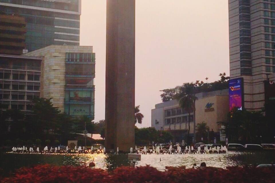 Uma estátua de uma pessoa com os braços levantados, um edifício alto e um corpo d'água com flores vermelhas ao primeiro plano.