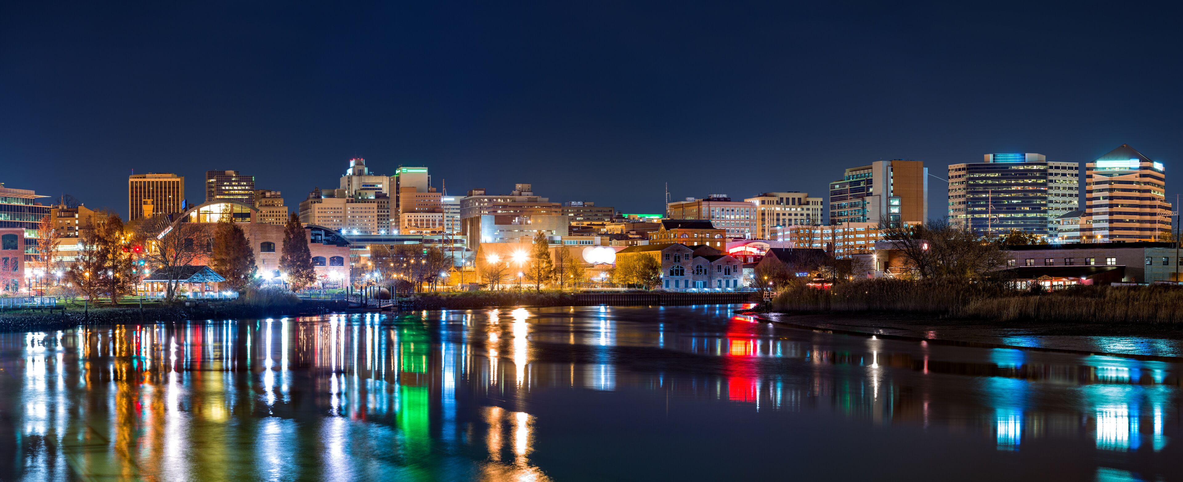 A nighttime cityscape with illuminated buildings reflecting on a calm river.