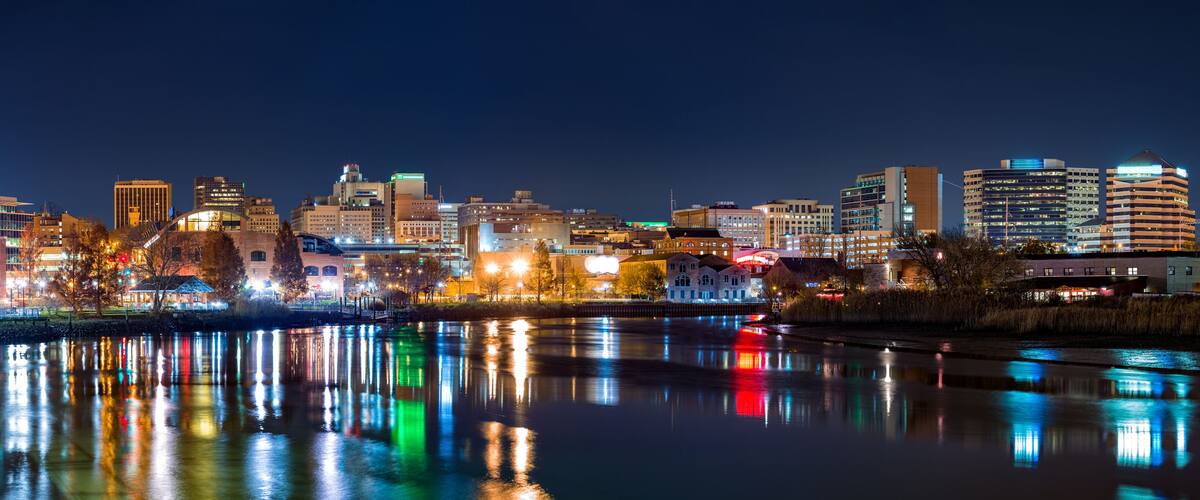 A nighttime cityscape with illuminated buildings reflecting on a calm river.
