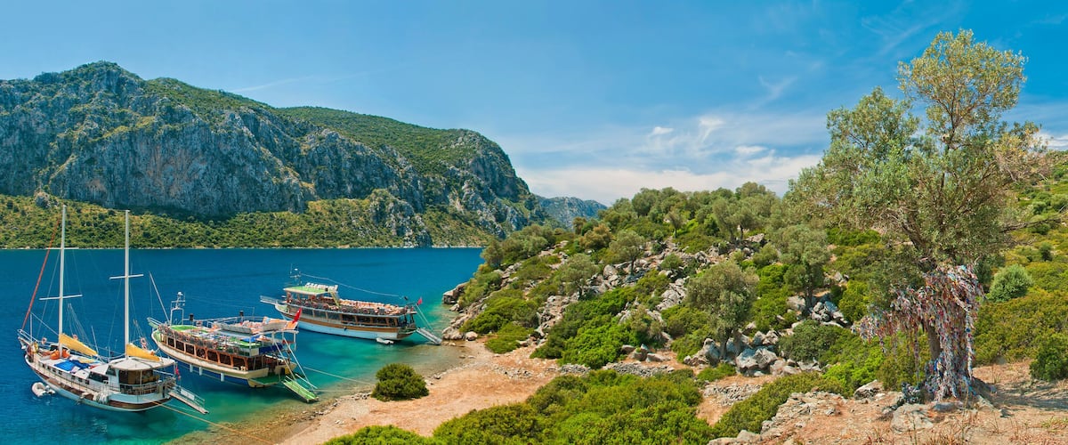 A coastal landscape with boats docked near a rocky shore, surrounded by lush greenery and mountains.