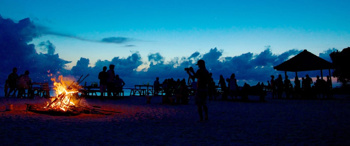 A beach scene at dusk with silhouettes of people around a campfire.