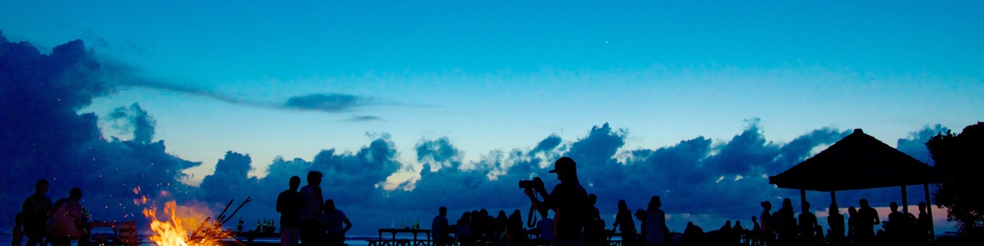 A beach scene at dusk with silhouettes of people around a campfire.