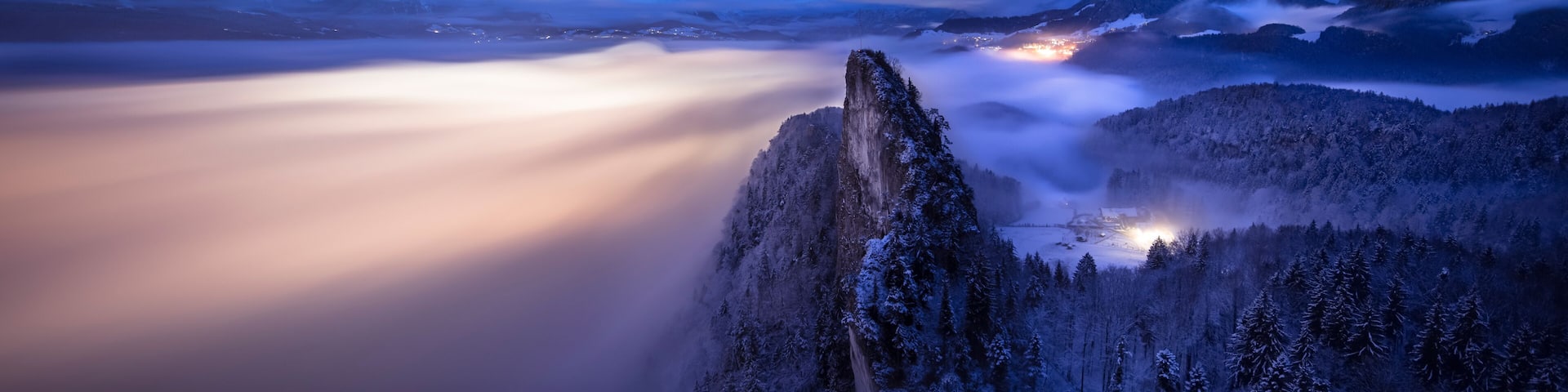 A snow-covered mountain peak, a forested valley, and a distant town illuminated by lights.