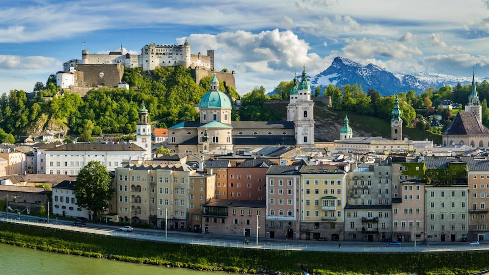 Paisagem urbana com um castelo imponente numa colina, um rio e uma cordilheira ao fundo.