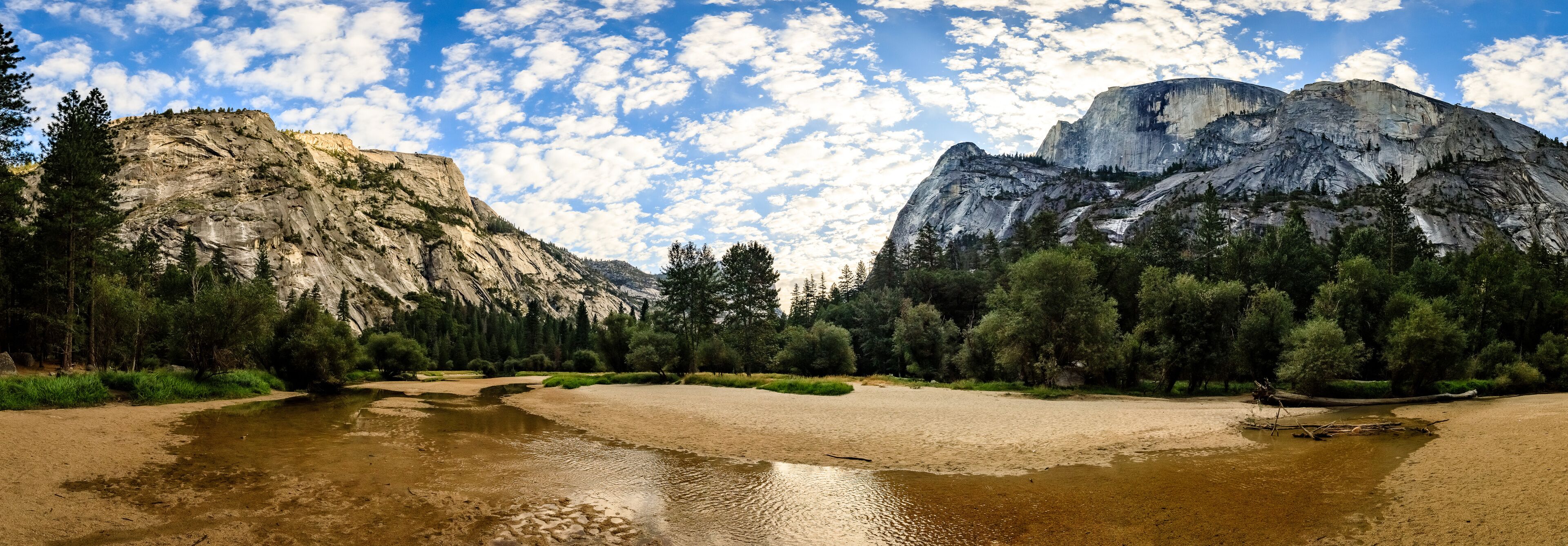 A river with a sandy bank, surrounded by dense forest and rocky mountains under a partly cloudy sky.