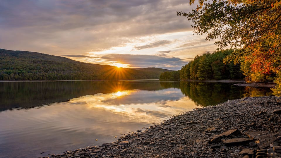 A calm lake with a sunset reflecting on the water, surrounded by forested hills.