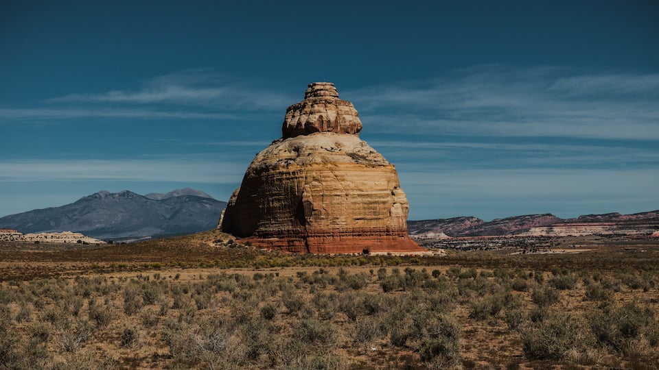 A large rock formation with a flat top and a smaller peak, surrounded by a sparse desert landscape and distant mountains.