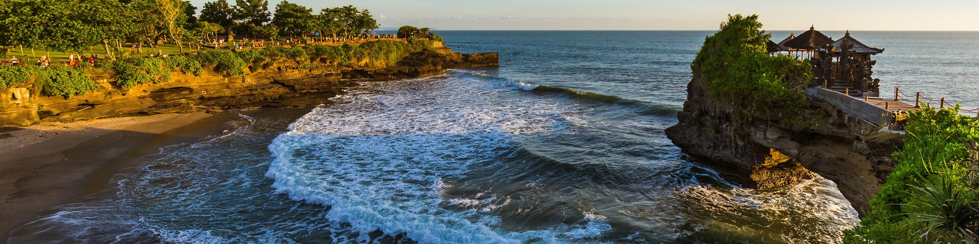 Paisagem costeira com ondas quebrando em uma praia arenosa, um afloramento rochoso com vegetação e uma estrutura de madeira à direita.