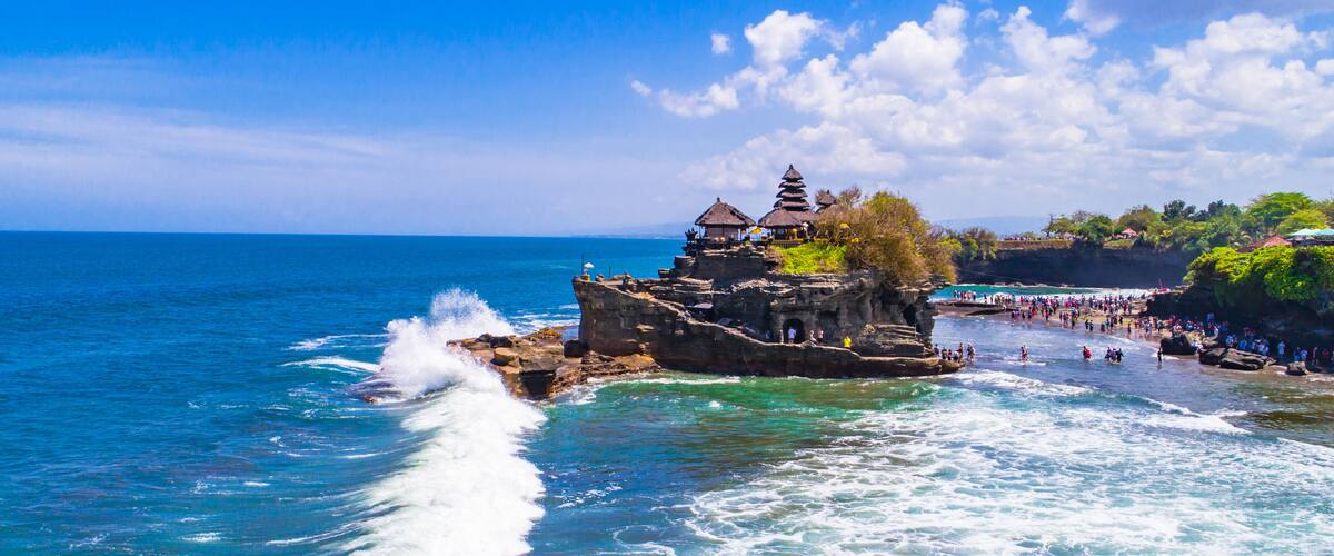 A rocky coastal area with clear blue waters, a small temple-like structure on a cliff, and a beach with people.