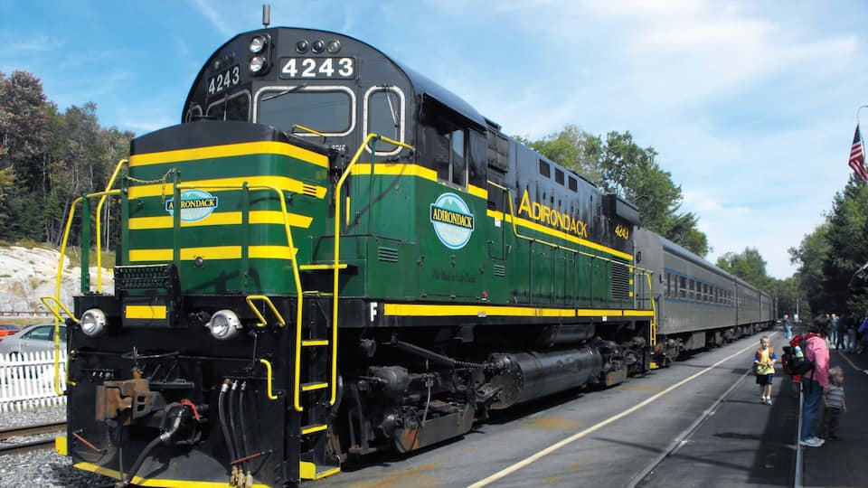 A green and yellow Adirondack train numbered 4243 on tracks with people and an American flag in the background.