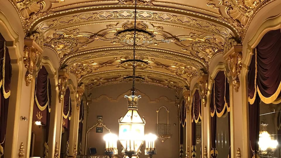 An ornate hallway with a grand chandelier, gold accents, and red curtains.