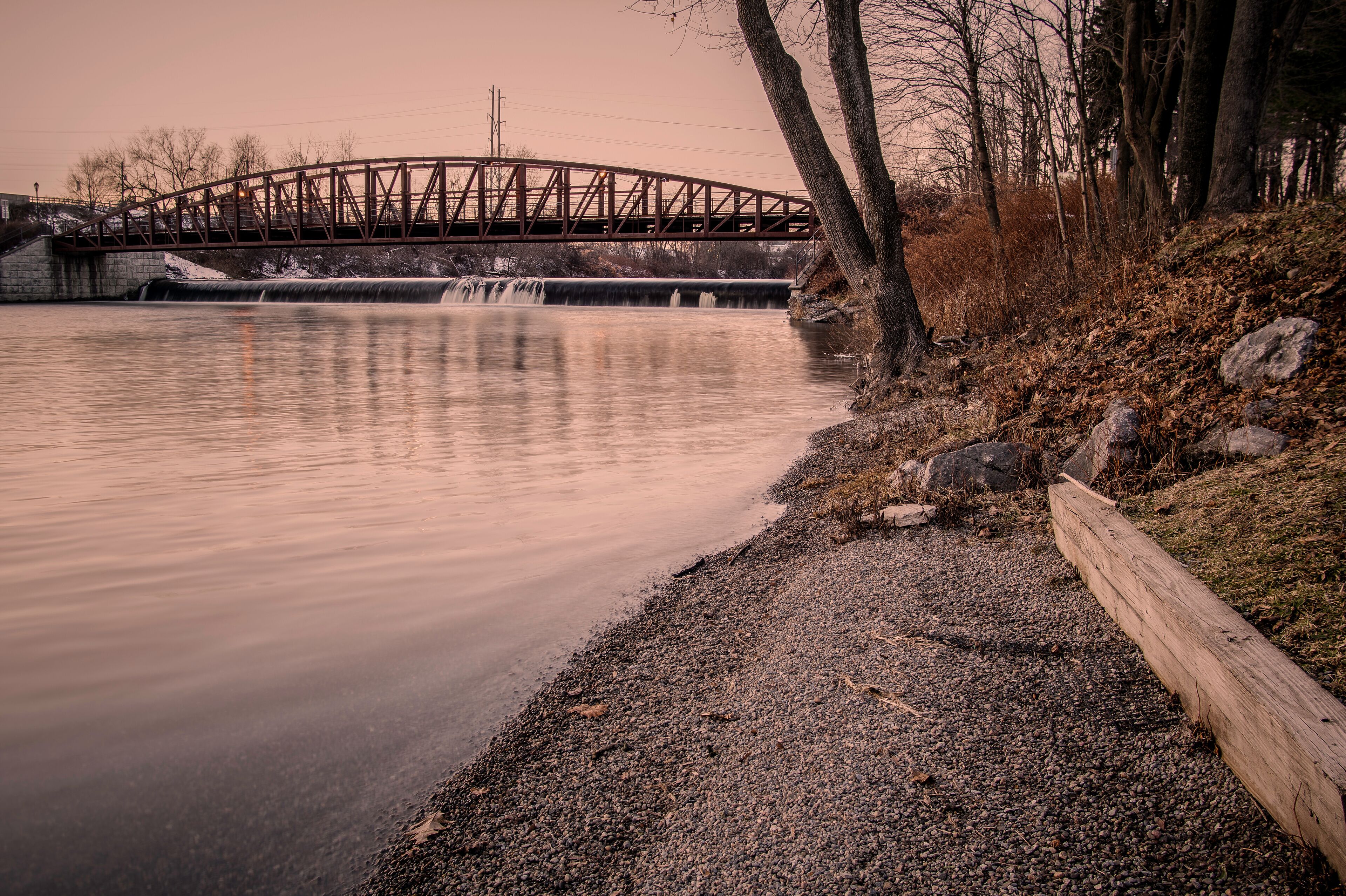 A calm river with a wooden walkway on the right, a metal bridge in the background, and leafless trees.