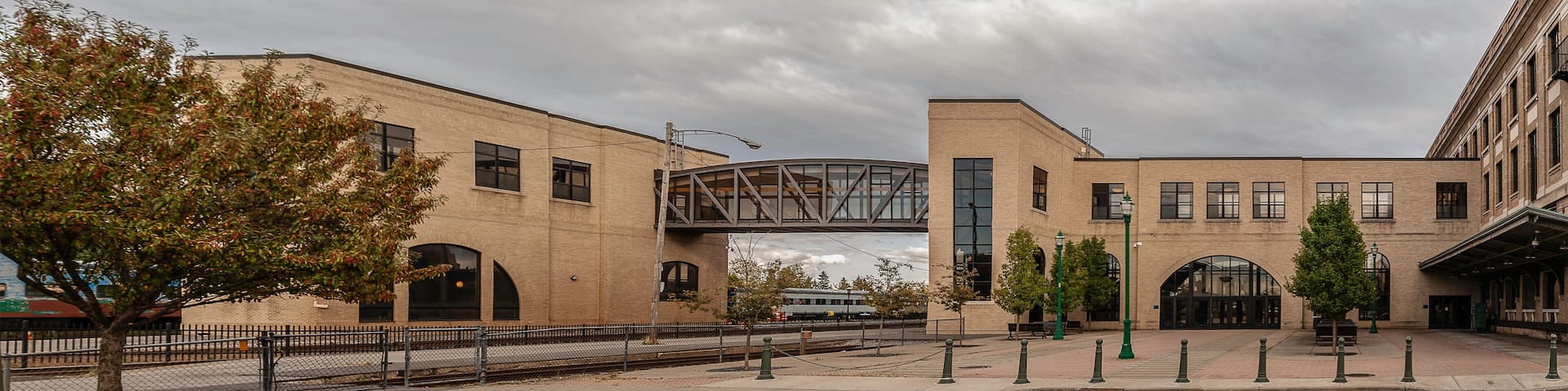 A train station with a pedestrian bridge, brick buildings, and trees.