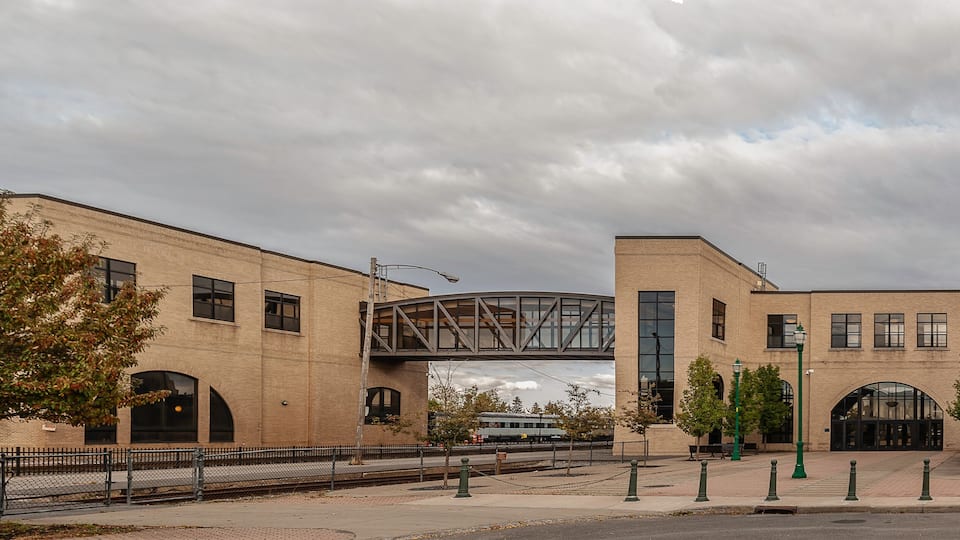 A train station with a pedestrian bridge, brick buildings, and trees.