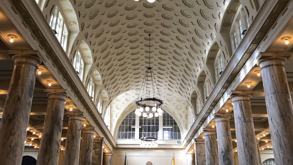 A grand hall with a high, patterned ceiling, large marble columns, and a central chandelier.