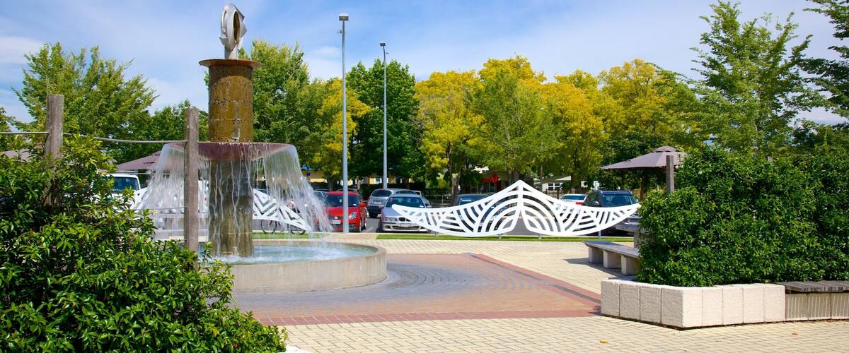 A fountain with a unique sculpture, surrounded by trees and a parking area.